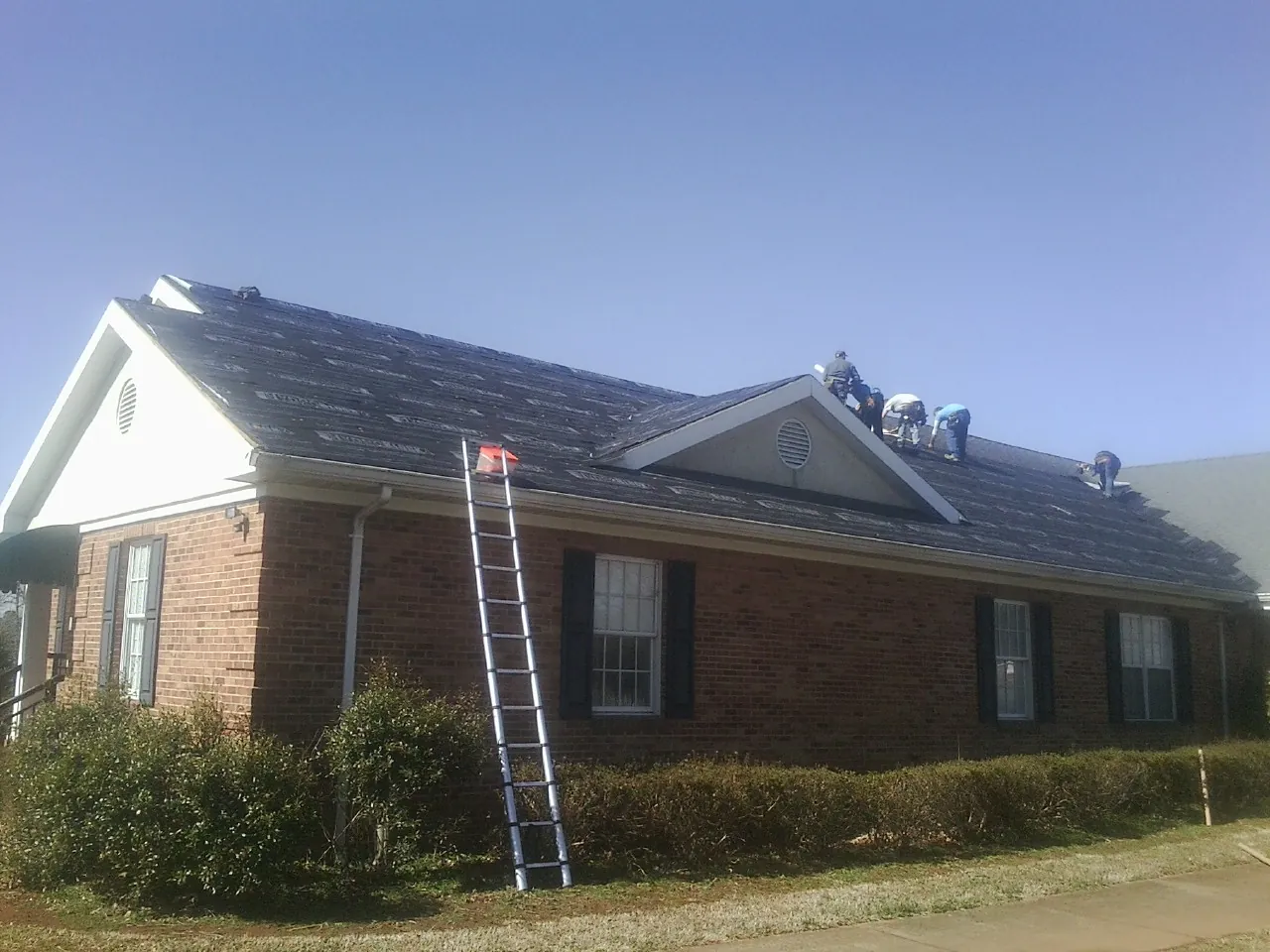 Crew tearing shingles off a brick ranch roof with a ladder set — Emergency Roof Repair work by our crew in Casnovia