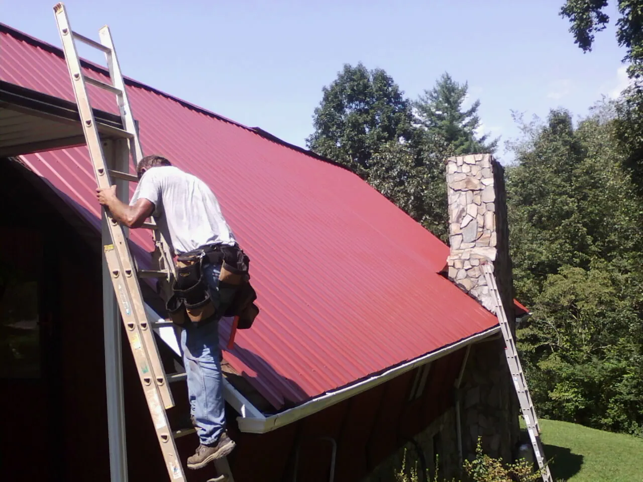Roofer on a ladder beside a red metal roof and stone chimney — Soffit & Fascia Repair by Roofing Brothers in Casnovia